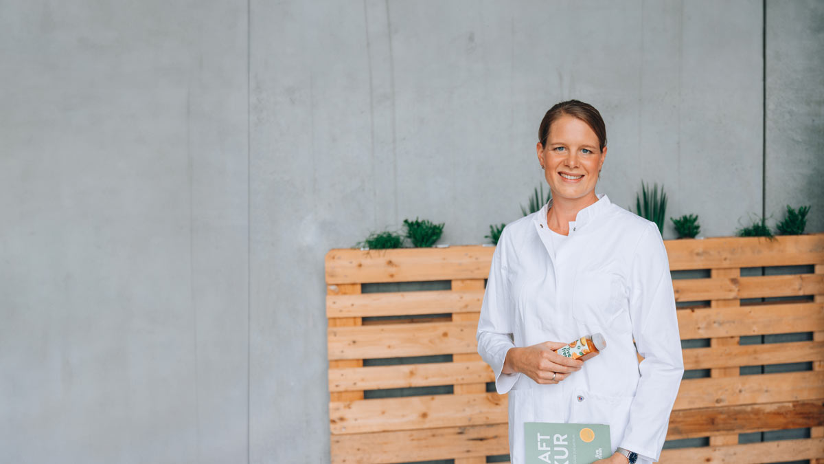 Dr. Ulrike Fischer, lächelnd in Kochuniform, hält eine Flasche LiveFresh Saft und ein Kochbuch, vor grauem Hintergrund mit Holzpalette und Pflanzen.