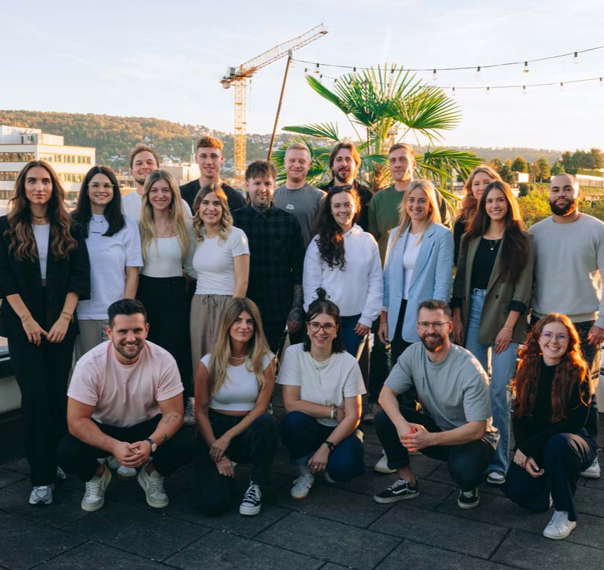 Gruppe von Menschen auf einer Dachterrasse bei Sonnenuntergang, mit Blick auf eine Stadtlandschaft mit Gebäuden, einem Kran und einer Palme.  Einige Personen hocken sich hin, die anderen stehen. Alle lächeln und posieren für ein gemeinsames Foto.