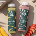 Zwei Flaschen Matcha Getränk von Live Fresh stehen nebeneinander auf hellem Untergrund, eine grüne Mango Matcha Latte und eine rosa Strawberry Matcha Latte. Daneben liegen frische Erdbeerscheiben und ein Stück Mango, die die fruchtigen Geschmacksrichtungen der Matcha Getränke visuell unterstreichen.