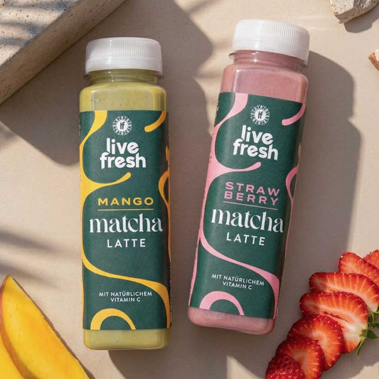 Zwei Flaschen Matcha Getränk von Live Fresh stehen nebeneinander auf hellem Untergrund, eine grüne Mango Matcha Latte und eine rosa Strawberry Matcha Latte. Daneben liegen frische Erdbeerscheiben und ein Stück Mango, die die fruchtigen Geschmacksrichtungen der Matcha Getränke visuell unterstreichen.