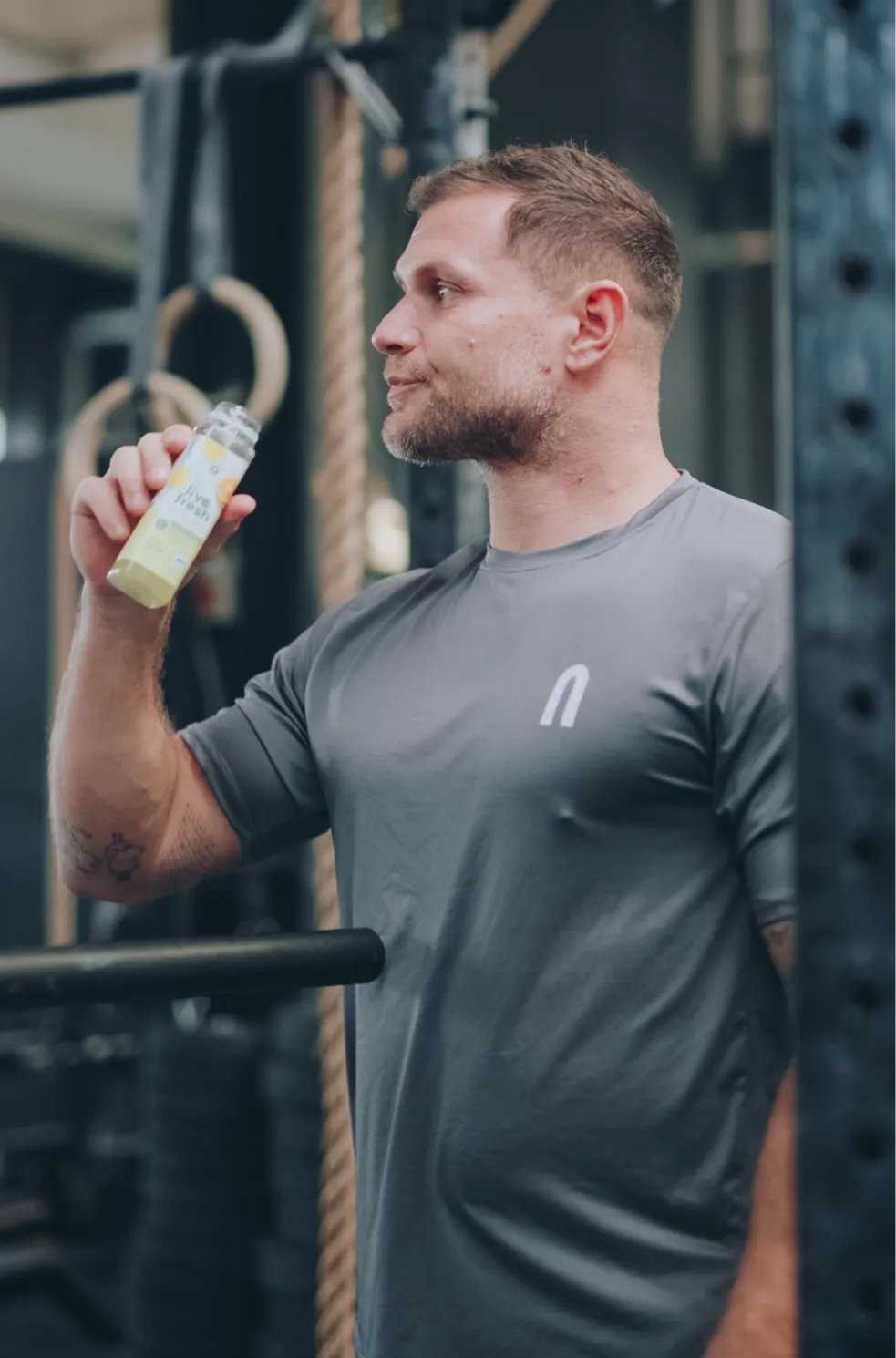 Mimi Krause, der ein graues T-Shirt trägt und seitlich steht. Er trinkt ein Saft von LiveFresh in einem Fitnessstudio, im Hintergrund sind Gymnastikringe und Trainingsgeräte zu sehen.