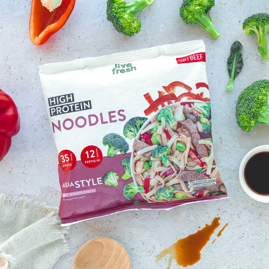 Das erste Bild zeigt eine Packung „Live Fresh High Protein Noodles“ in der Geschmacksrichtung Asia Style mit Rindfleisch. Die Verpackung hebt den Proteingehalt von 35 g und die Zubereitungszeit von 12 Minuten hervor. Im Hintergrund sind frische Zutaten wie Brokkoli, Paprika, und Sojasauce zu sehen, was die asiatische Inspiration unterstreicht.