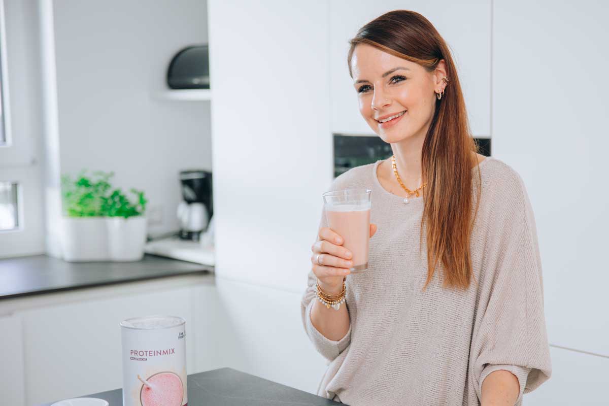 Eine junge Frau, die lächelnd ein Glas mit einem rosa Proteinshake in der Hand hält. Sie steht in einer hellen, modernen Küche und trägt ein beiges, locker sitzendes Oberteil. Im Vordergrund steht eine Dose Proteinmix, die das Produkt darstellt. Die freundliche und einladende Atmosphäre vermittelt ein Gefühl von Leichtigkeit und Gesundheit, wobei der Fokus auf einem aktiven und ernährungsbewussten Lebensstil liegt.