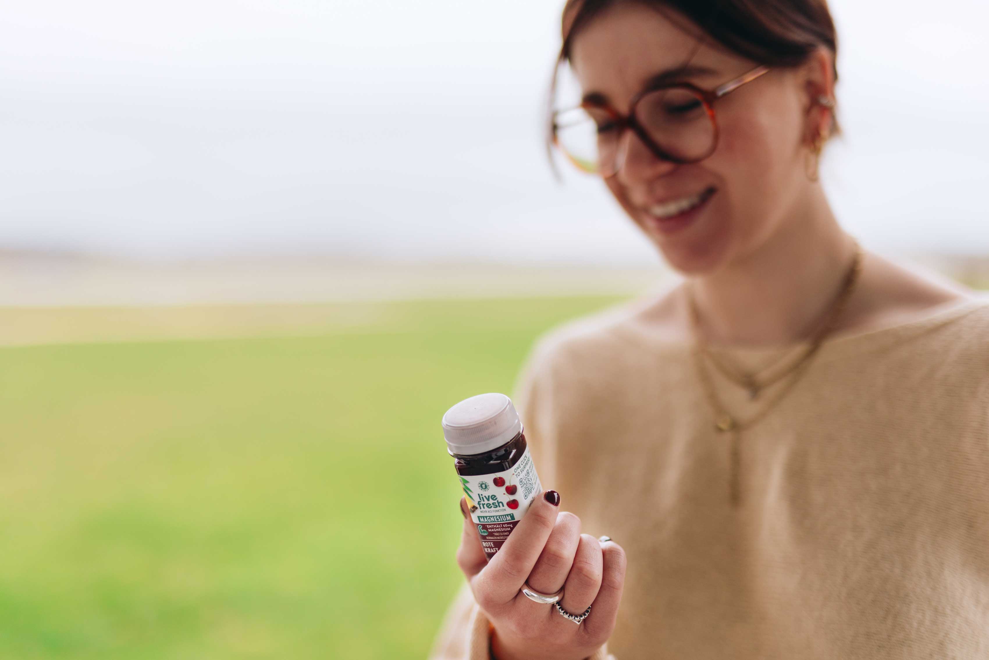 Eine Person hält lächelnd einen Shot namens Rote Kraft von LiveFresh, im Hintergrund ist verschwommen eine grüne Wiese zu erkennen. 