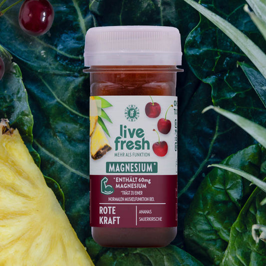 LiveFresh Shot „Rote Kraft“ mit Magnesium für Muskelfunktion. Dunkelrot gefärbt, liegt er auf Grünkohl, umgeben von Ananas, Sauerkirschen und Blättern.