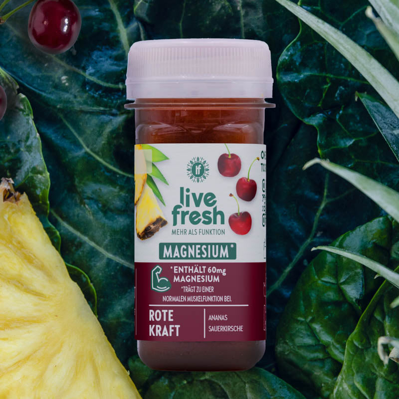 LiveFresh Shot „Rote Kraft“ mit Magnesium für Muskelfunktion. Dunkelrot gefärbt, liegt er auf Grünkohl, umgeben von Ananas, Sauerkirschen und Blättern.