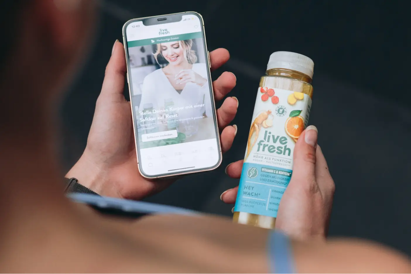 Person hält ein Smartphone mit der LiveFresh-App geöffnet in der einen Hand und einen LiveFresh-Saft in der anderen Hand.