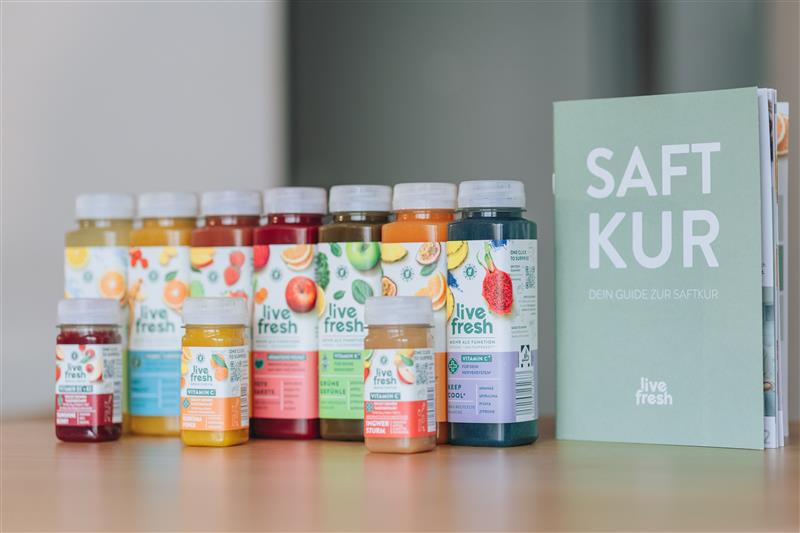 Sieben verschiedene Säfte von LiveFresh und drei Shots auf hellem Holzuntergrund. Daneben steht eine grüne Broschüre mit der Aufschrift "Saftkur".