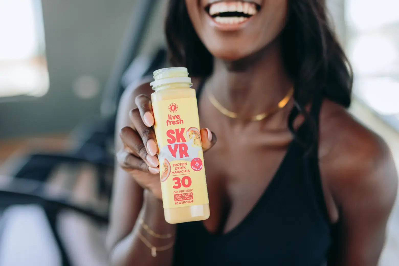 Frau im Fitnessstudio hält eine geöffnete Flasche LiveFresh SKYR Protein Drink Maracuja mit 30 Gramm Protein in die Kamera.