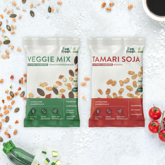 Zwei Packungen von 'Live Fresh' Snacks, Veggie Mix und Tamari Soja, nebeneinander auf einer hellen Steinoberfläche mit gestreuten Sojabohnen und Gemüsestücken.