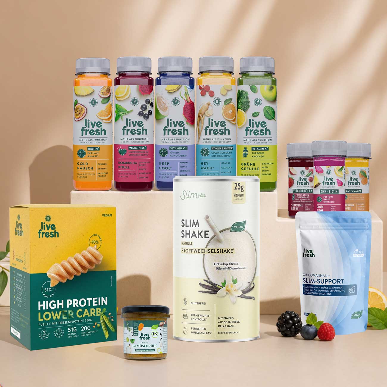 Eine Auswahl von Live Fresh Produkten ist auf beigefarbenen Podesten arrangiert. Zu sehen sind mehrere farbige Functional Drinks, Proteinpulver, High Protein Low Carb Fusilli, Nahrungsergänzungsmittel, Glucomannan Slim Support sowie ein Glas Gemüsebrühe, dekoriert mit frischem Obst, Beeren und Kräutern vor einem hellen Hintergrund.