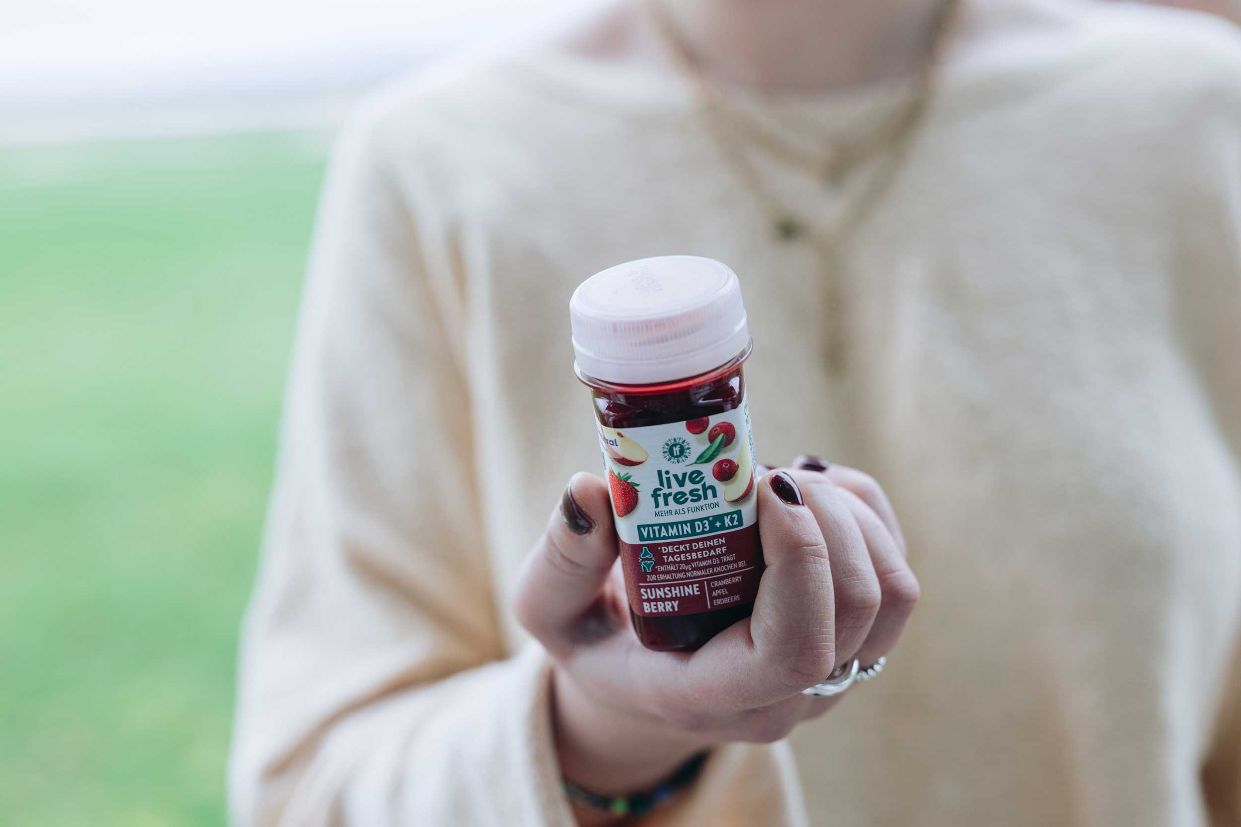 Person hält einen  Vitamin-D3+K2 Shot namens Sunshine Berry von LiveFresh in der Hand, im Hintergrund ist verschwommen eine Wiese zu erkennen. 