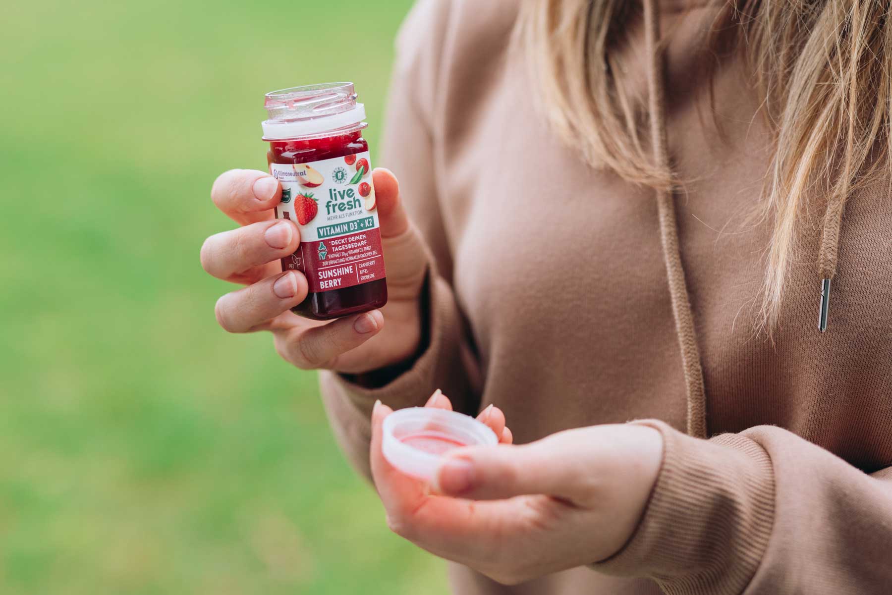 Person hält einen geöffneten Vitamin-D3+K2 Shot namens Sunshine Berry der Marke LiveFresh in der Hand, im Hintergrund ist ein Feld zu erkennen. 