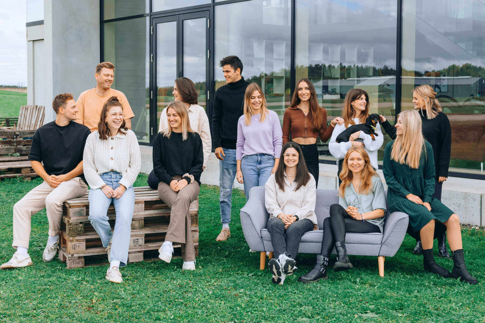 Eine Gruppe von zwölf jungen Menschen sitzt und steht entspannt vor einem modernen Bürogebäude mit großen Glasfenstern. Sie lachen und unterhalten sich, während eine Person einen kleinen schwarzen Hund hält, der von einer anderen gestreichelt wird. Die Gruppe ist leger gekleidet, einige sitzen auf Paletten, andere auf einem grauen Sofa, mit einer grünen Wiese im Vordergrund und einem ländlichen Hintergrund.