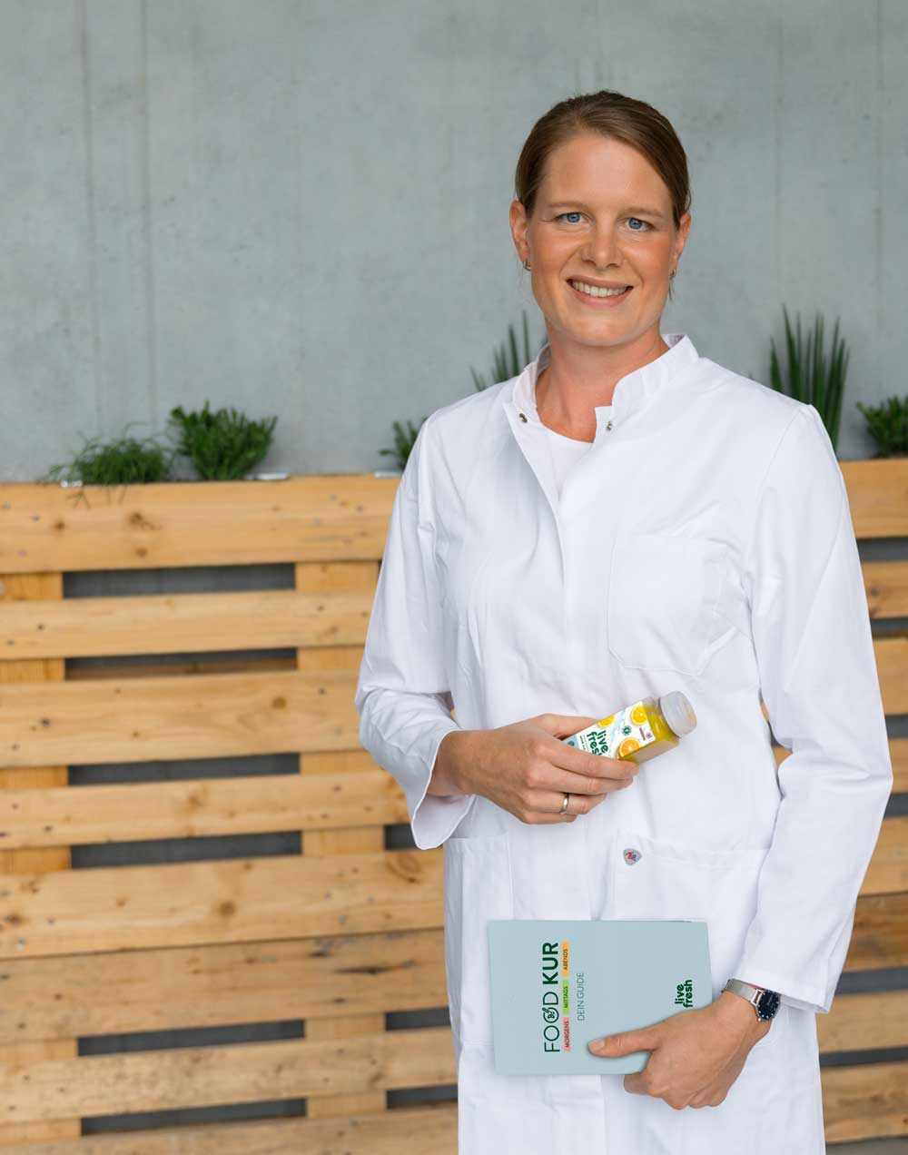 Eine lächelnde Frau mit braunen, zusammengebundenen Haaren in einem weißen Kittel hält eine Flasche Saft von LiveFresh in der einen Hand und ein Buch mit der Aufschrift "FOOD KUR" in der anderen Hand. Im Hintergrund stehen Pflanzen auf Holzpaletten vor einer grauen Wand.