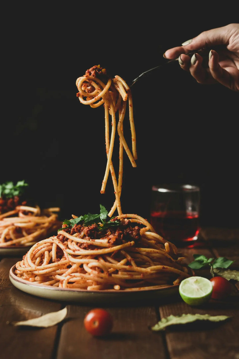 Gabel hebt lange Spaghetti mit Bolognese über einen Teller mit Pasta auf einem Holztisch. Im Hintergrund stehen Kräuter, Tomaten und ein Glas Rotwein vor dunklem Hintergrund.