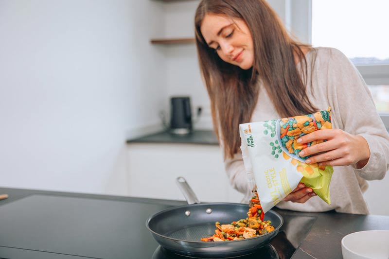 Eine Frau in einer modernen Küche, die eine „Live Fresh“-Verpackung mit der Aufschrift „Bowl Exotic Style“ in eine Pfanne gibt. In der Pfanne befinden sich bereits bunte Gemüsestücke, die als Teil eines gesunden, veganen Gerichts zubereitet werden. Die Frau lächelt leicht und scheint konzentriert bei der Zubereitung zu sein. 