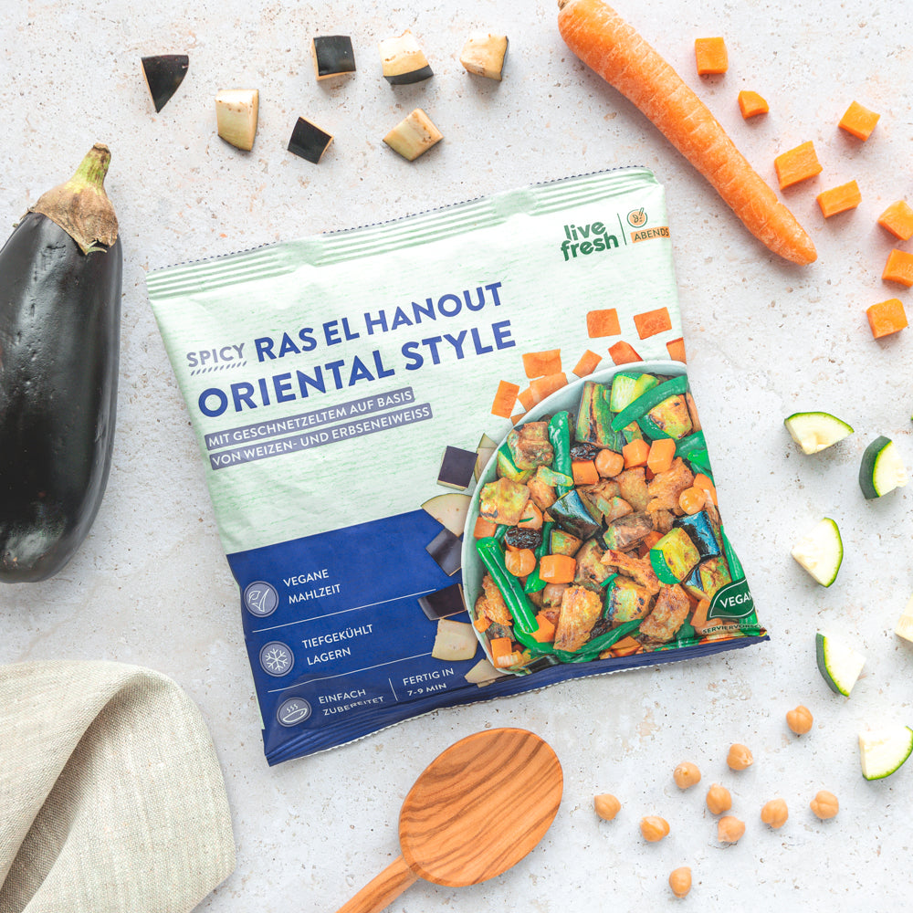 Eine Packung „Spicy Ras El Hanout Oriental Style“ von Live Fresh, umgeben von frischen Zutaten wie Auberginen, Karottenstückchen, Zucchini und Kichererbsen. Daneben liegt eine Holzkelle, die das Bild einer schnellen, gesunden und veganen Mahlzeit für den Abend abrundet. Die Verpackung betont, dass die Mahlzeit tiefgekühlt ist und in 7-9 Minuten zubereitet werden kann.