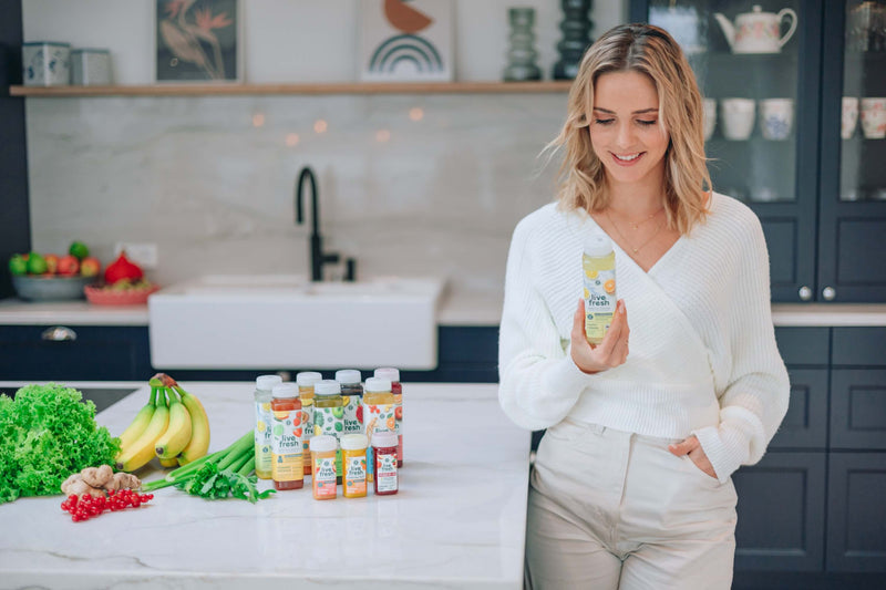 Blonde Frau in weißem Pullover und heller Hose steht lächelnd in einer dunklen Küche. Links neben ihr stehen verschiedene Säfte von LiveFresh, außerdem Obst und Gemüse wie Sellerie. Sie sieht auf den Saft in ihrer Hand. 